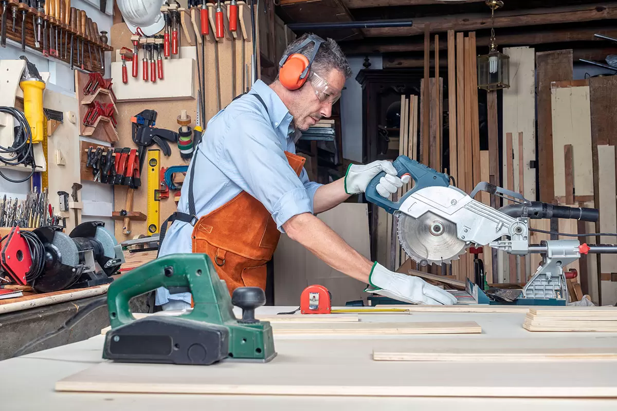 A man wearing safety gear works with a miter saw in a woodshop. He is focused on cutting a wooden board. The workshop is filled with tools, including a green power tool on the table and various hand tools hanging on the wall.
