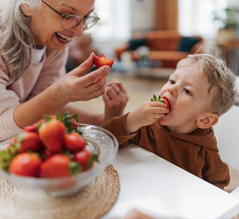 An older woman and a young boy sit at a table, smiling and eating fresh strawberries together. A glass bowl filled with strawberries is in the foreground.