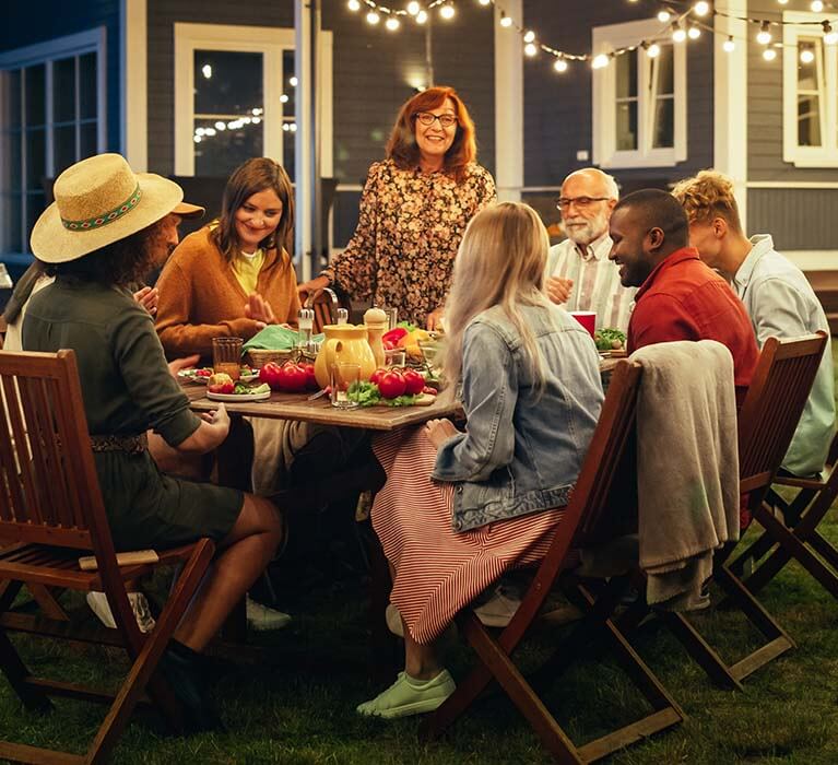 A group of people enjoying an outdoor dinner together at a table in a backyard at night, smiling and talking under string lights, with plates of food in front of them.