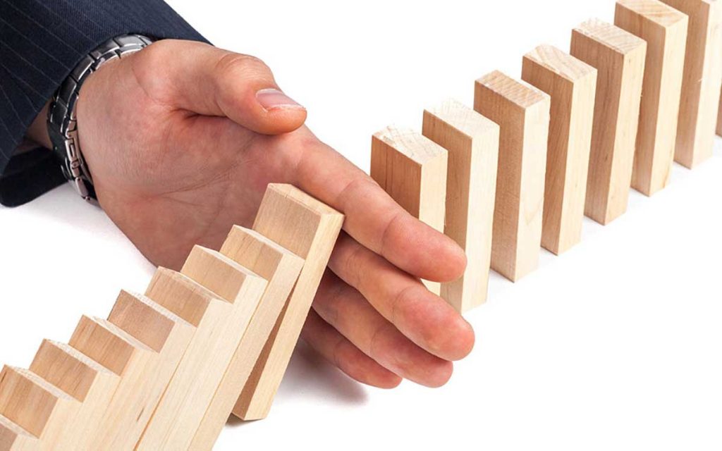 A hand in a business suit interrupts a row of falling wooden dominoes, stopping the chain reaction on a white background.