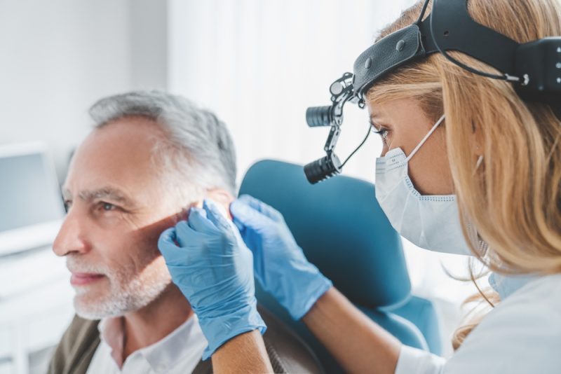 A doctor wearing a headlamp and mask examines the ear of an older man with gray hair, who is sitting in a medical office chair. The doctor is using gloved hands for the examination.
