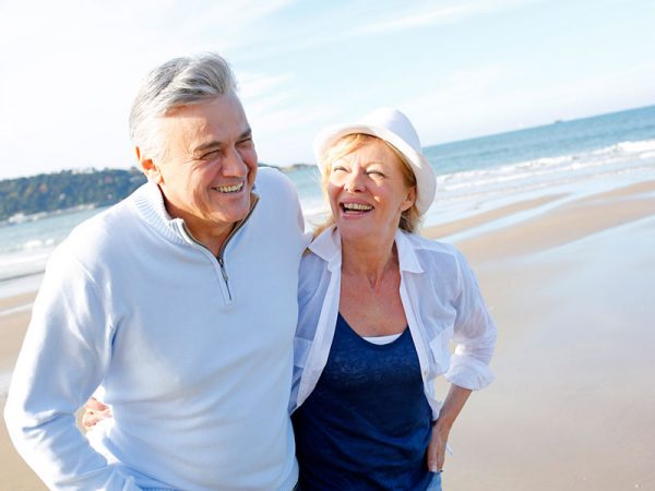 An older couple smiles and walks arm in arm along a sandy beach, with the ocean and sky in the background. They appear happy and relaxed, dressed in light, casual clothing.