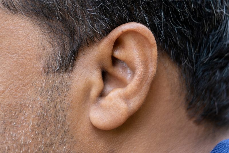 Close-up view of a persons ear and part of their face, showing short dark hair with some gray strands and textured skin.