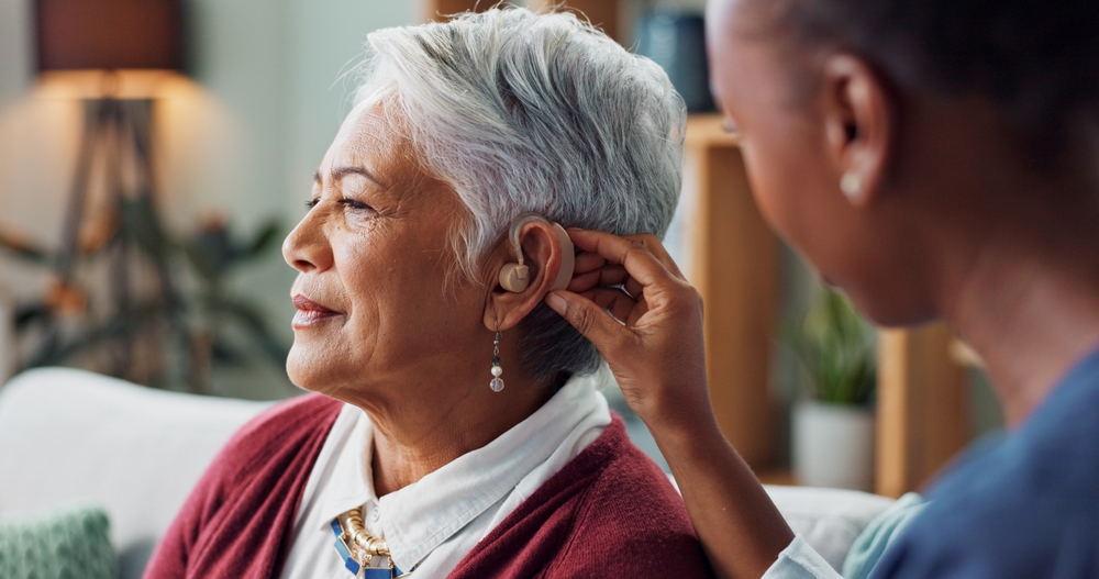 A smiling older woman with short gray hair sits as another person helps adjust her hearing aid. They are indoors, and the scene appears warm and supportive.