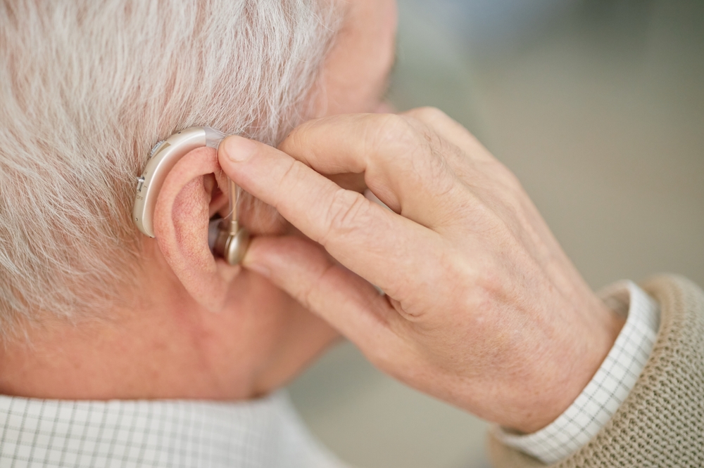 Close-up of an older adult adjusting a behind-the-ear hearing aid on their left ear with their right hand. The person has gray hair and is wearing a checked shirt and beige sweater.