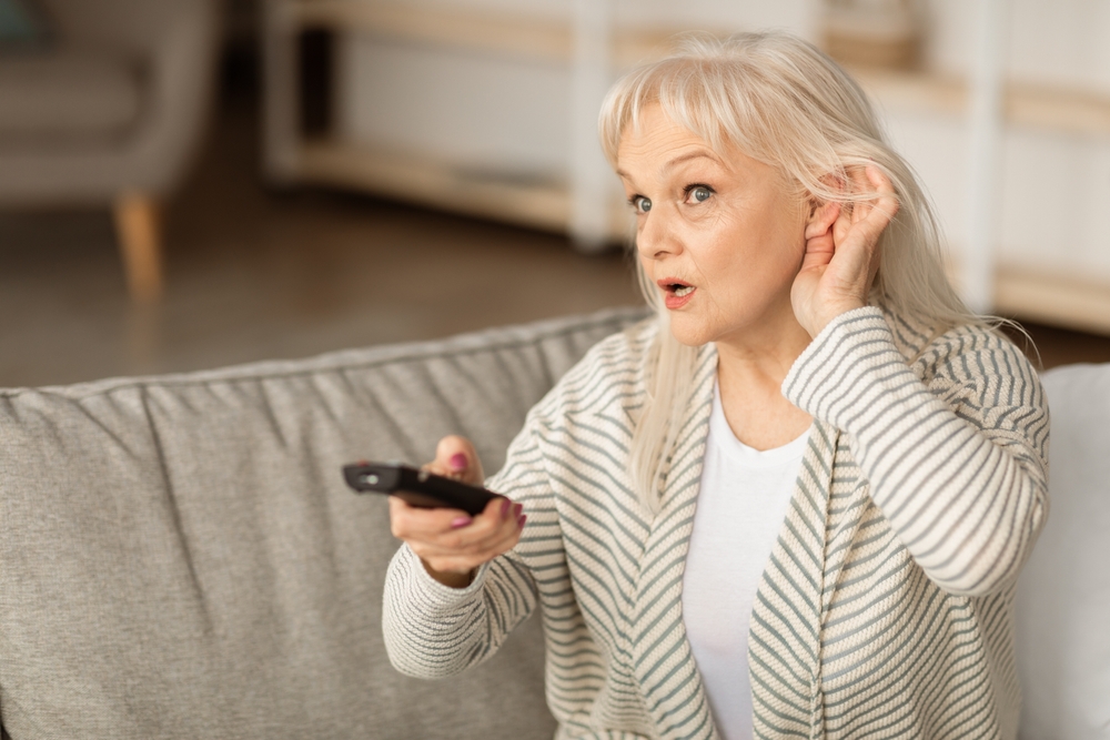 An older woman sitting on a couch holds a TV remote in one hand and cups her ear with the other, appearing to have difficulty hearing. She looks attentive and slightly concerned.