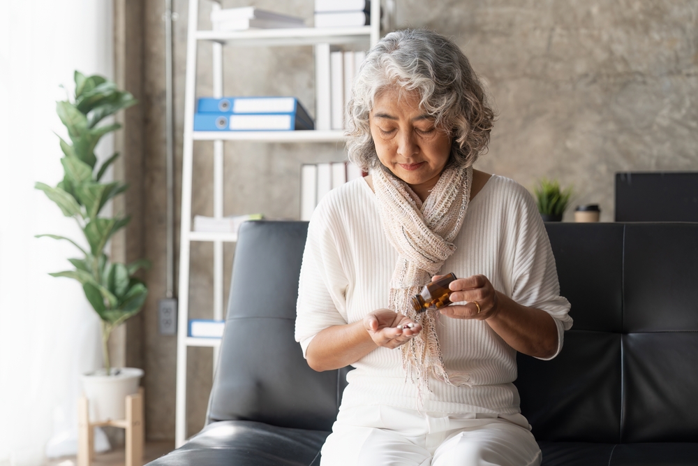 An older woman with gray hair sits on a couch, pouring pills from a brown bottle into her hand. She wears a white top and scarf, with bookshelves and a plant in the background.