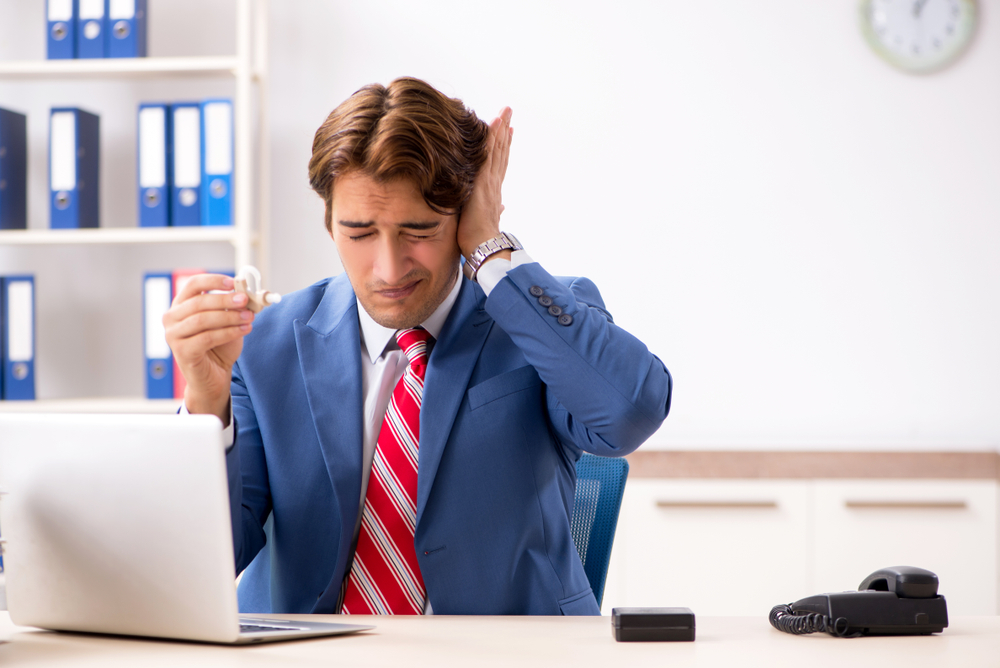 A man in a suit sits at a desk with a laptop, holding his ear in discomfort and holding a small object, possibly an earplug, with a pained expression. Office items are visible in the background.