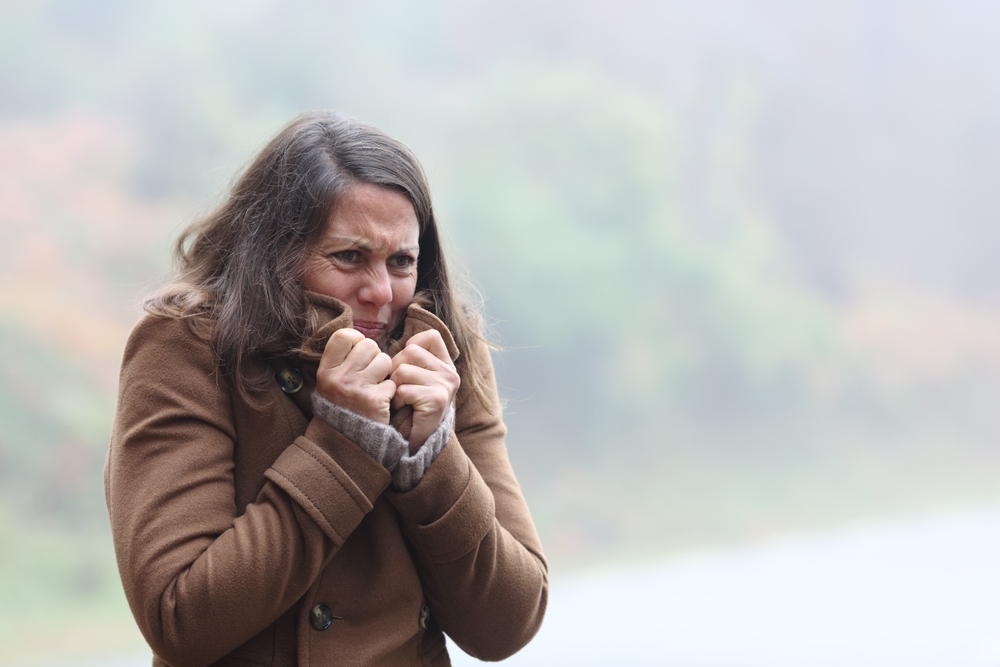 A woman wearing a brown coat stands outdoors, clutching her collar and shivering from the cold, with a blurred background of greenery and mist.