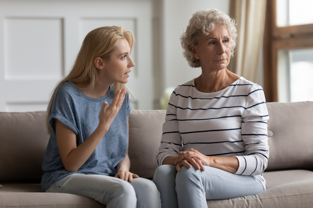 A young woman, looking upset, gestures while talking to an older woman with gray hair who sits beside her on a couch, looking away with a serious expression.