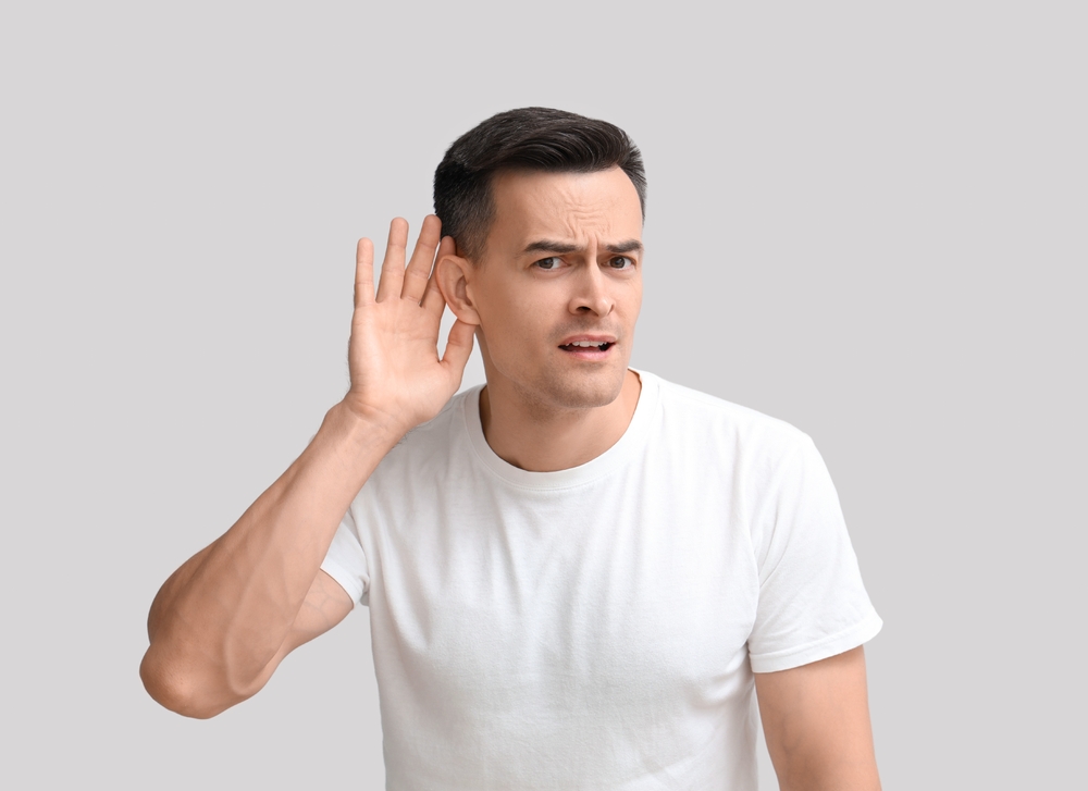 A man in a white t-shirt holds his hand to his ear and looks confused, as if trying to hear something better, against a plain light gray background.