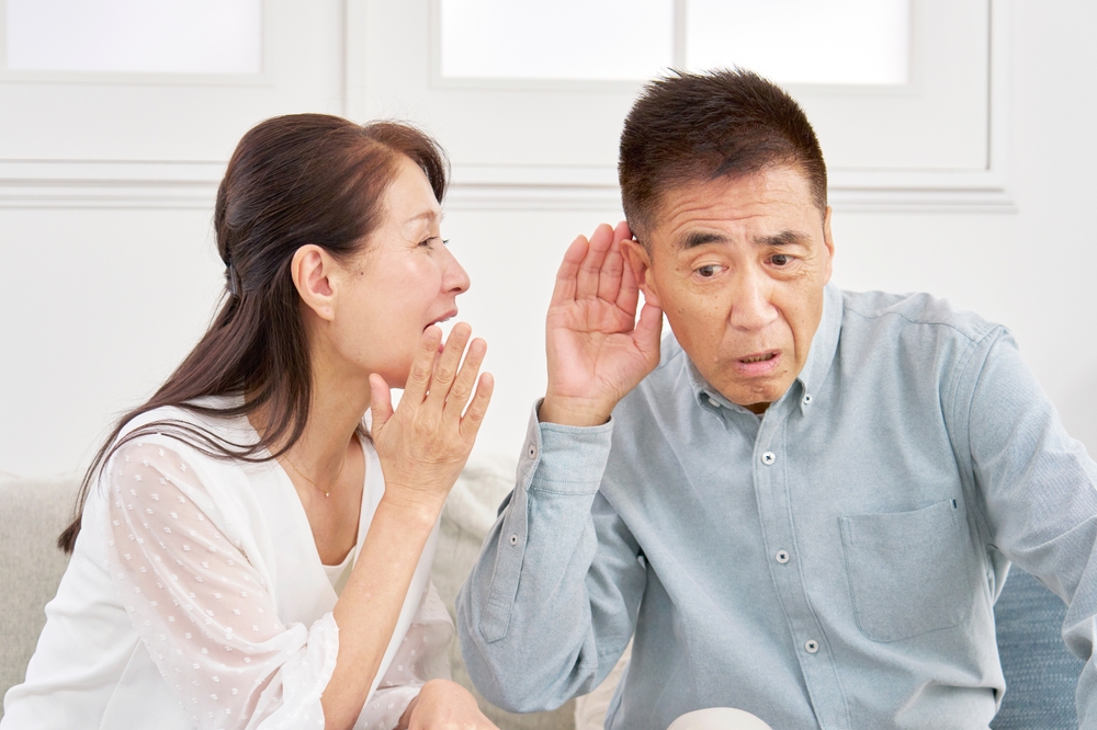 A woman is whispering to a man who is holding his hand to his ear, appearing to have difficulty hearing her. They are sitting indoors, both wearing light-colored clothing.