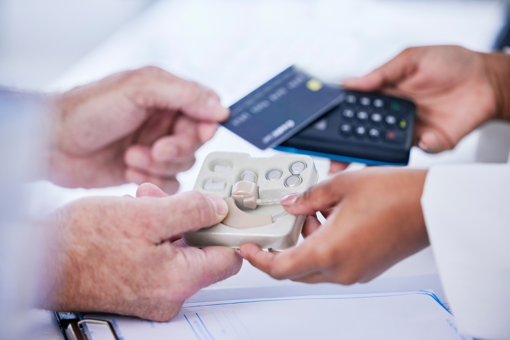 Two people exchange a hearing aid device in packaging while one person holds a credit card and another holds a payment terminal, suggesting a purchase or transaction in a medical setting.