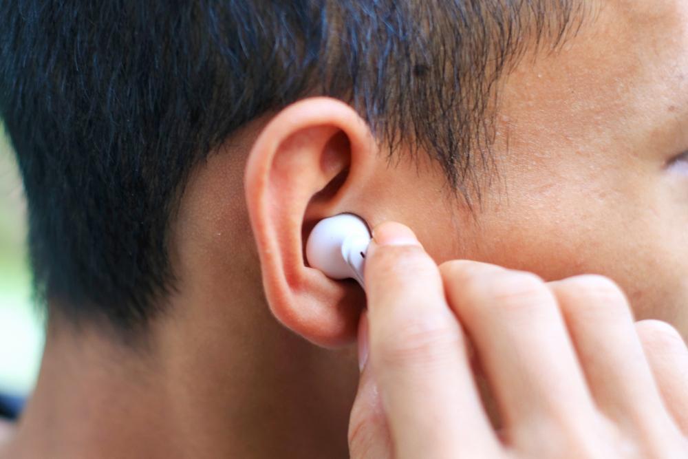 A person is inserting a white wireless earbud into their right ear, with their hand touching the earbud. The image focuses on the ear and the earbud, capturing the action up close.