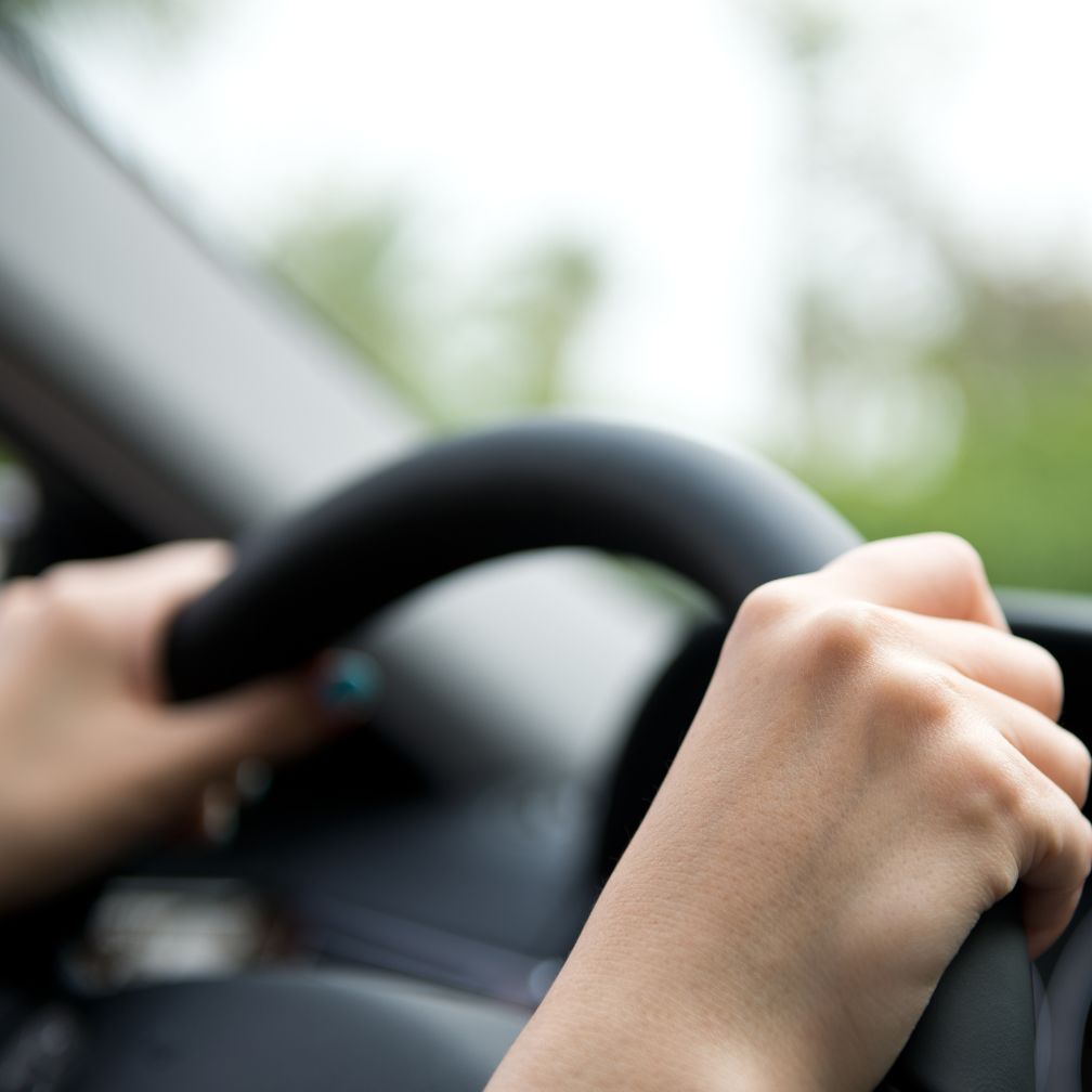 Close-up of a person’s hands gripping a car steering wheel, with an out-of-focus view through the windshield in the background.