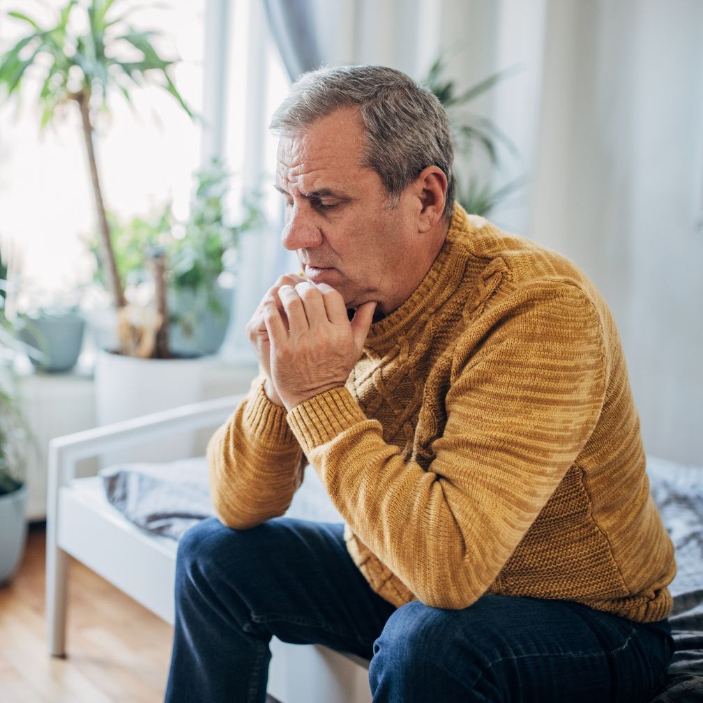 An older man in a mustard sweater sits on a bed, looking thoughtful and concerned, with hands clasped under his chin in a bright room with plants.