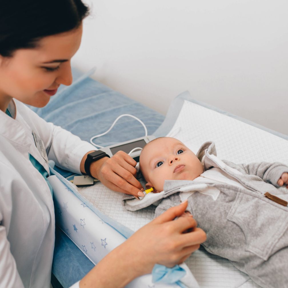 A healthcare professional is performing a hearing test on a baby lying on an exam table. The baby is wearing a grey outfit and looking up while the professional carefully places equipment on the babys ear.