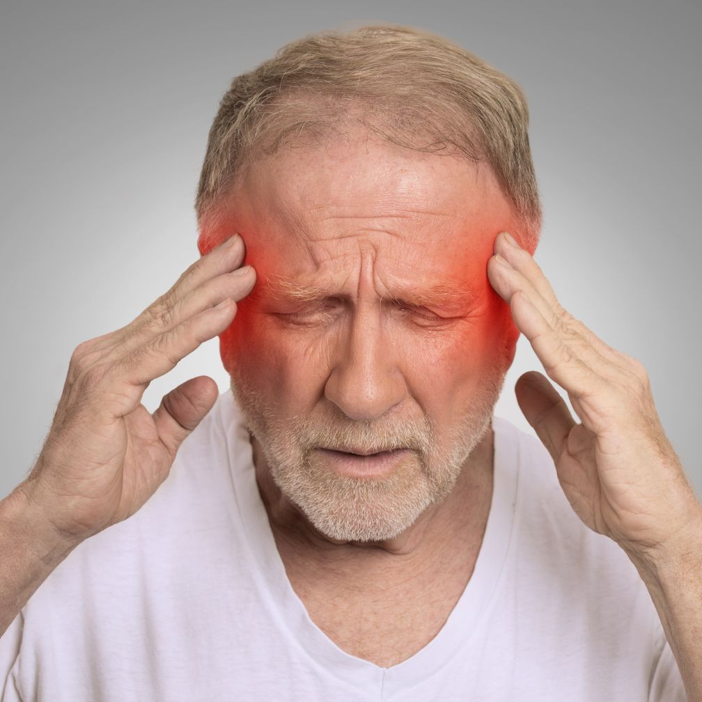 An older man touches his temples with both hands, eyes closed, appearing in pain. Red shading around his temples suggests a headache or migraine.