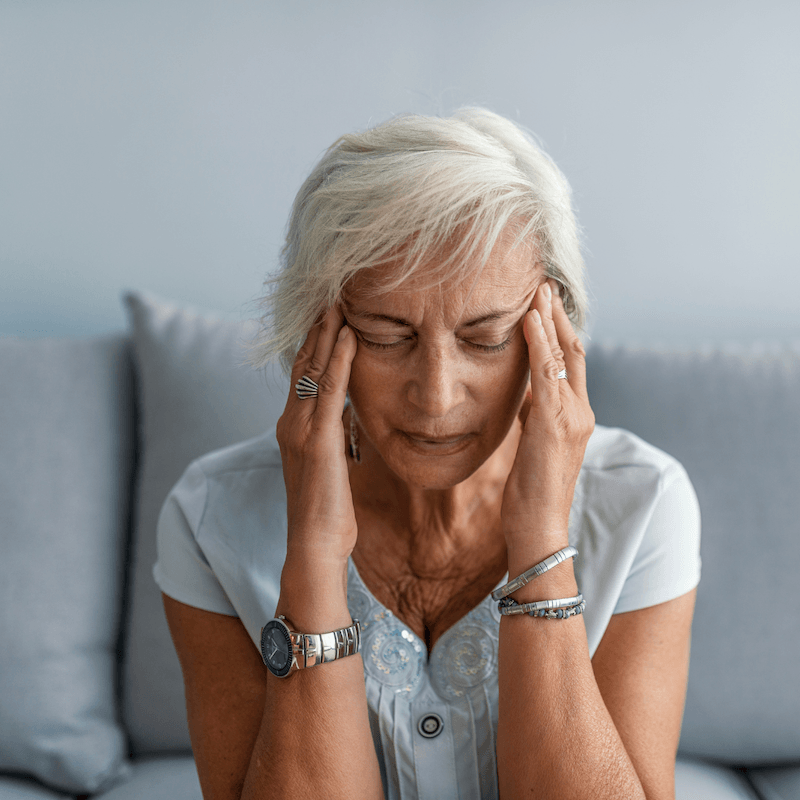 Older woman with short white hair sits on a couch, eyes closed, pressing her temples with both hands as if experiencing a headache or stress, wearing a light blue shirt and silver jewelry.