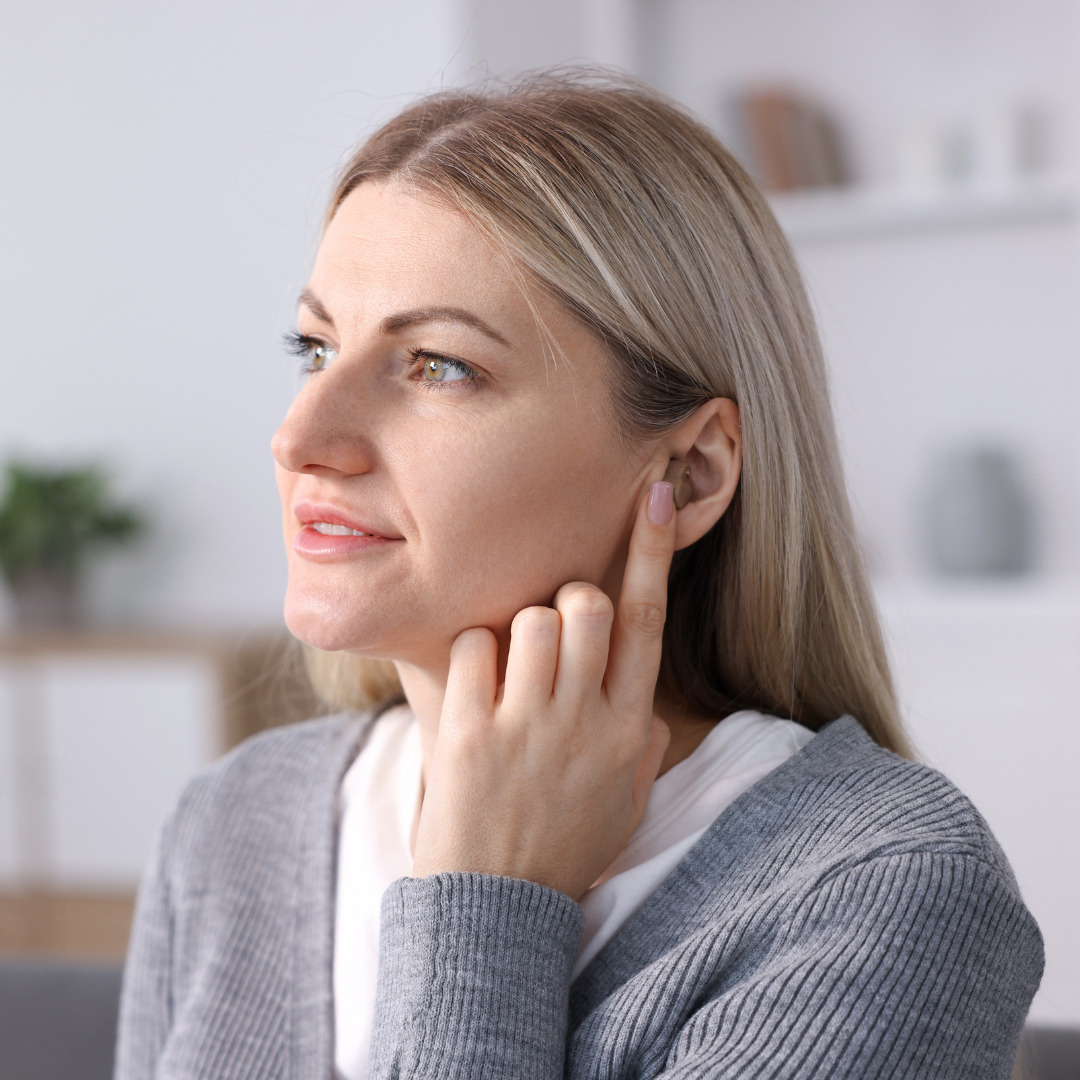 A woman with blonde hair touches her ear with her index finger while looking to the side, wearing a gray cardigan indoors.